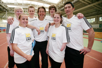 Eine Gruppe von sieben jungen Menschen, darunter auch Menschen mit Behinderung, steht lächelnd in einem Hallensportstadion. Sie tragen weiße T-Shirts mit dem Logo der "Bundesliga"-Liga und schwarzen Shorts oder Hosen. Ein junger Mann legt den Arm um die Schulter einer jungen Frau. Im Hintergrund ist die rote Laufbahn der Halle zu sehen.