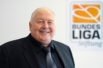 Ein Mann mit kurzem, grauem Haar lächelt freundlich in die Kamera. Im Hintergrund ist das Logo der DFL Stiftung mit dem Schriftzug "Bundesliga Stiftung" zu sehen.