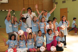 Eine Gruppe von Kindern hält Fußballbälle in die Höhe, umgeben von zwei erwachsenen Betreuerinnen in einem Turnsaal.