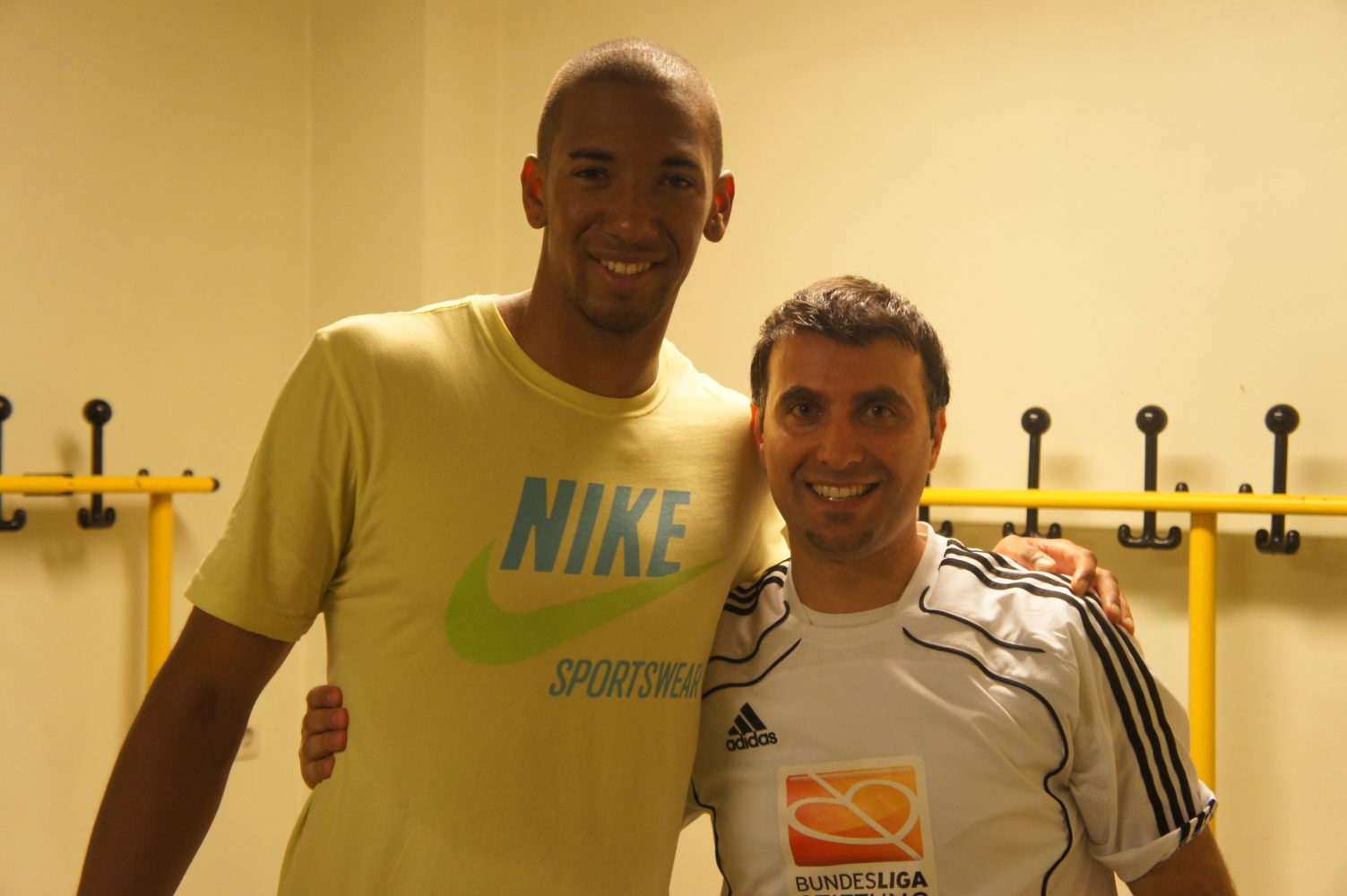 Zwei Männer, beide lächelnd, posieren für ein Foto. Der Mann links trägt ein hellgelbes T-Shirt mit dem Aufdruck "NIKE" und "SPORTSWEAR". Der Mann rechts trägt ein weißes T-Shirt mit dem Adidas-Logo und einem orangefarbenen Designelement mit dem Schriftzug "Bundesliga". Im Hintergrund sind Haken an einer hellen Wand zu sehen.