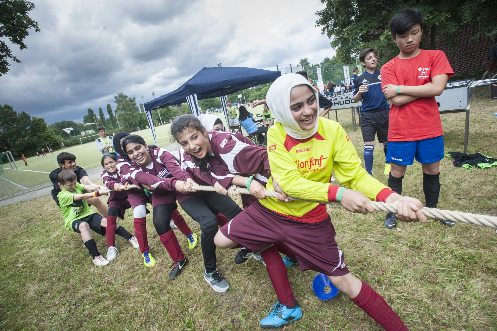 Eine Gruppe Jugendlicher zieht beim IKW Cup, einem Fußballturnier in Berlin, am Seil beim Tauziehen. Einige tragen rote T-Shirts, andere dunkelrote Sportkleidung mit gelben Akzenten und eine Person trägt ein gelbes T-Shirt mit Kopftuch. Im Hintergrund sind weitere Personen und Zelte zu sehen.