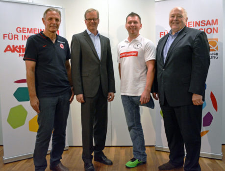 Vier Männer posieren für ein Foto vor einem Banner mit bunten Fußballmotiven. Von links: Ein Mann in rotem Poloshirt, ein Mann im Anzug, ein Mann in weißem T-Shirt mit Werbeaufdruck und ein weiterer Mann im Anzug.