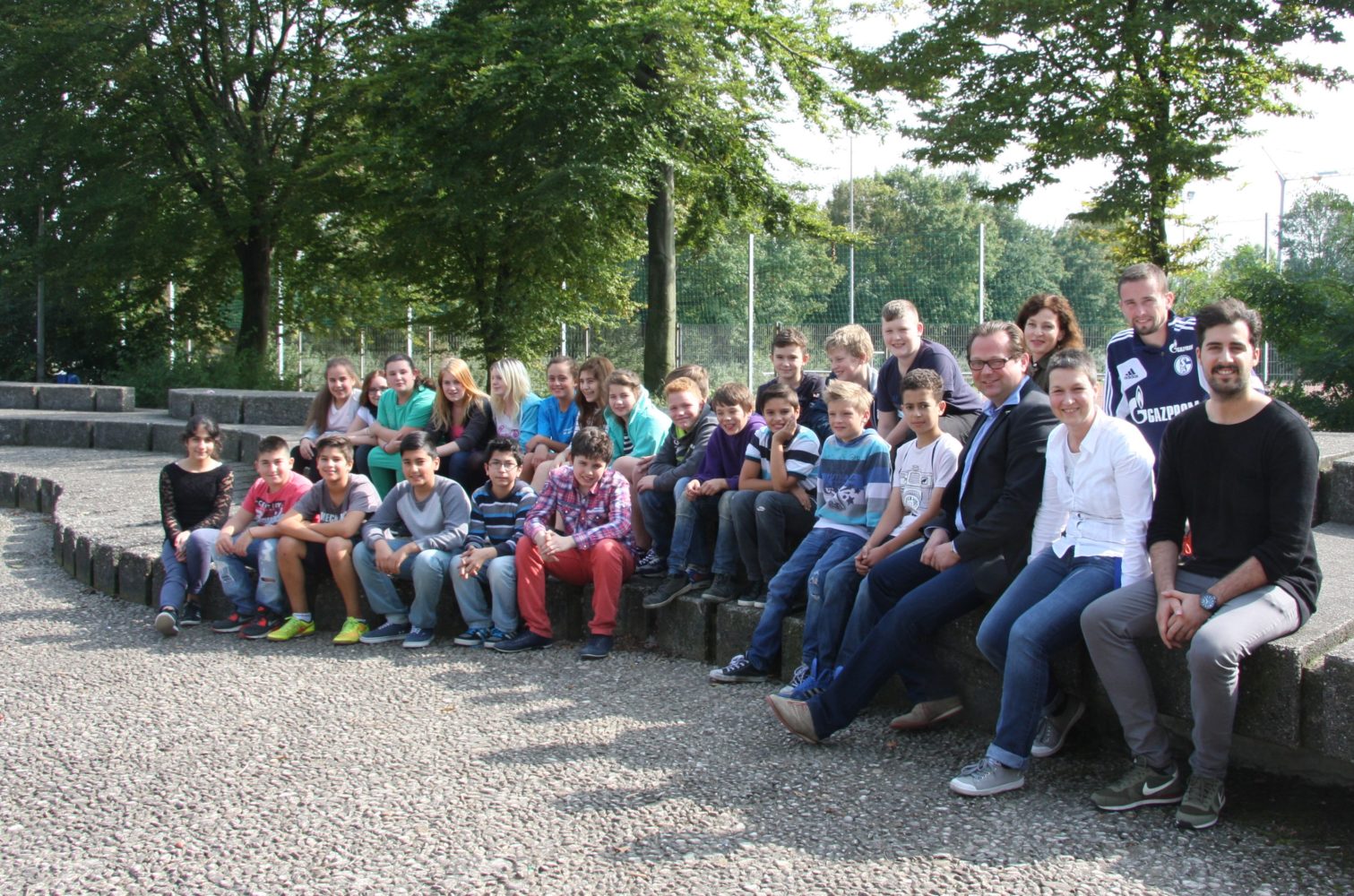 Eine Gruppe von Schülerinnen und Schülern sitzt und steht auf einer Stufe vor einer Mauer. Im Hintergrund sind Bäume und ein Zaun zu sehen.