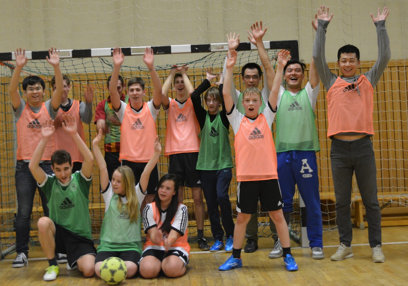 Eine Gruppe von Jugendlichen und jungen Erwachsenen in grünen und orangen Trainingsshirts jubelt gemeinsam mit erhobenen Armen in einer Sporthalle. Im Vordergrund knien einige Teilnehmer um einen Fußball.
