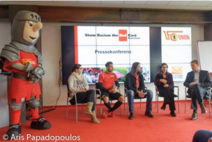 Eine Gruppe von fünf Personen sitzt auf einer Bühne vor einem Banner mit der Aufschrift "Show Racism the Red Card". Links steht eine große, rote Ritterfigur, das Maskottchen der DFL Stiftung.