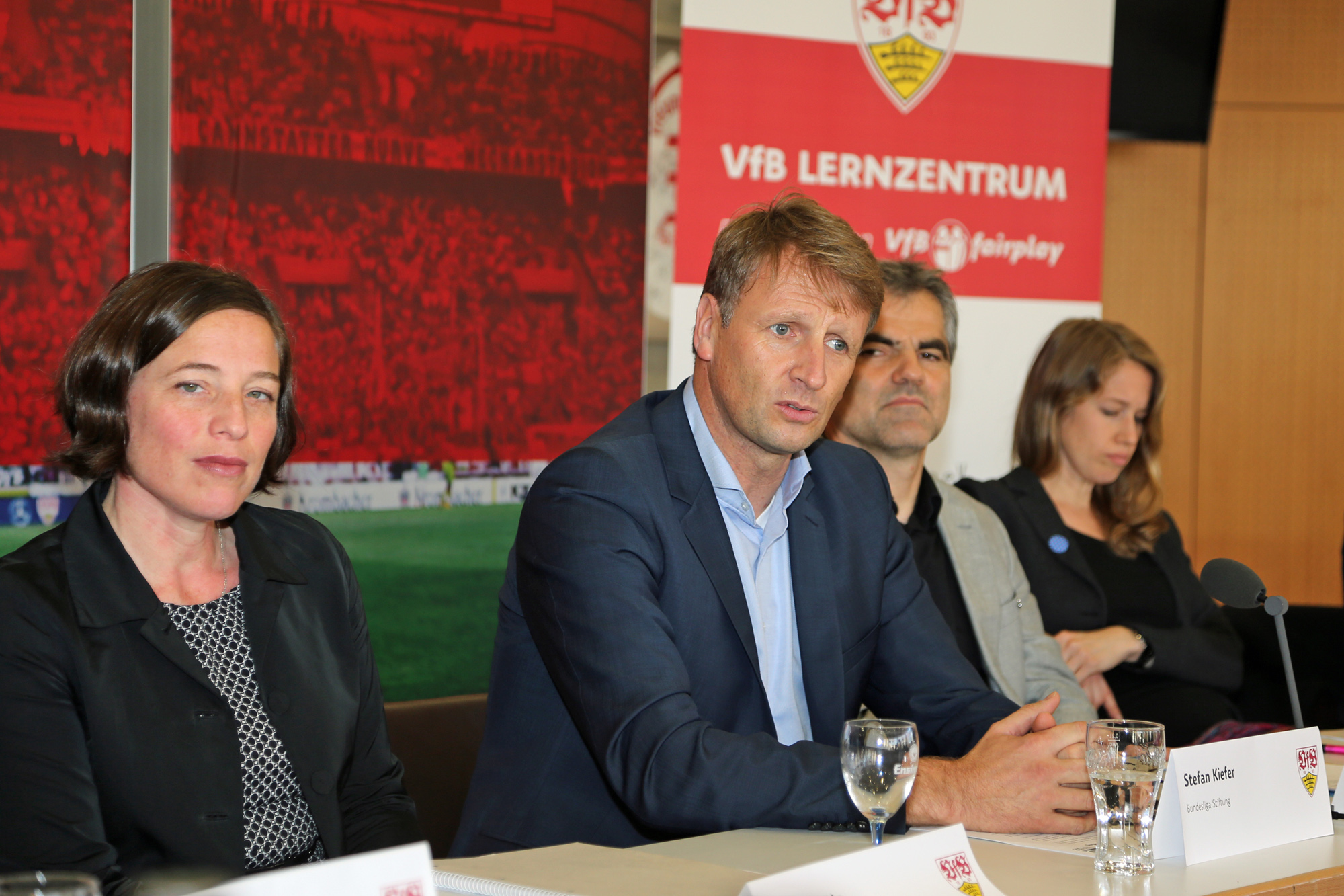 Eine Gruppe von Menschen sitzt an einem Tisch in einem Konferenzraum, vermutlich bei einer Veranstaltung der DFL Stiftung im VFB Lernzentrum. Im Vordergrund ist ein Mann mit blonden Haaren in einem dunklen Blazer zu sehen, der den Blick auf das Publikum richtet. Neben ihm sitzen weitere Personen, darunter eine Frau mit dunklen Haaren und eine weitere Frau mit langen, roten Haaren. Im Hintergrund ist ein Banner mit der Aufschrift "VFB LERNZENTRUM" und dem Logo von "fairplay" zu sehen.