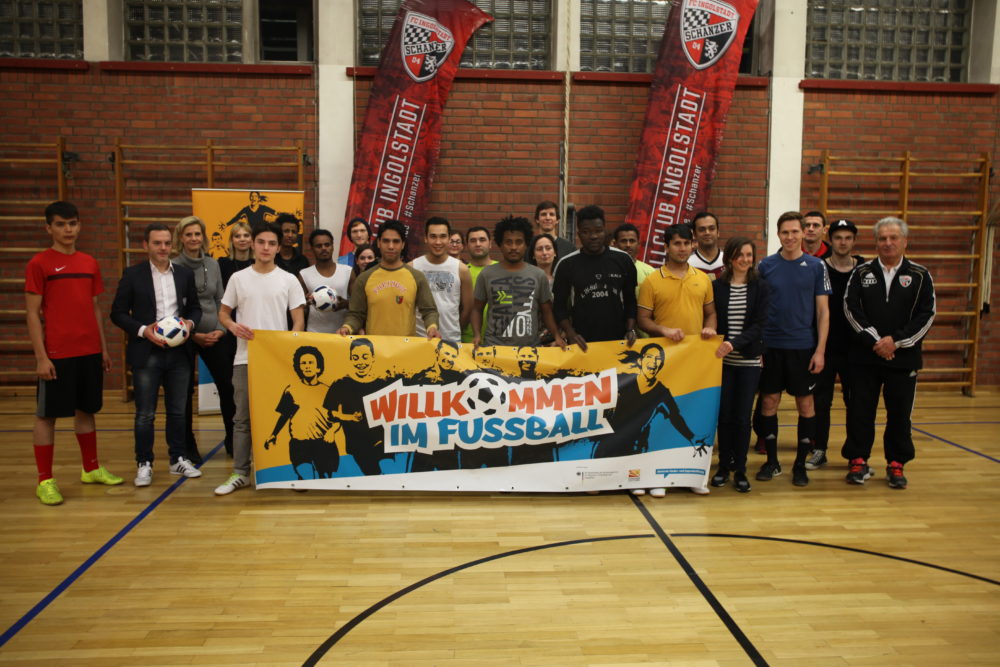 Eine Gruppe von Jugendlichen und Erwachsenen steht in einer Turnhalle vor einer Wand mit Bannern des FC Ingolstadt. Sie halten ein großes Banner mit der Aufschrift "Willkommen im Fußball".