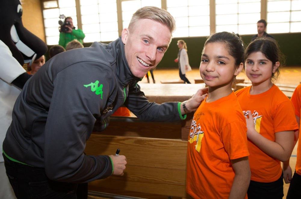 Ein lächelnder Fußballspieler kniet vor zwei jungen Mädchen und legt ihr freundschaftlich die Hand auf die Schulter. Alle tragen orangefarbene T-Shirts mit einem Logo. Im Hintergrund sind weitere Personen und eine Sporthalle erkennbar.