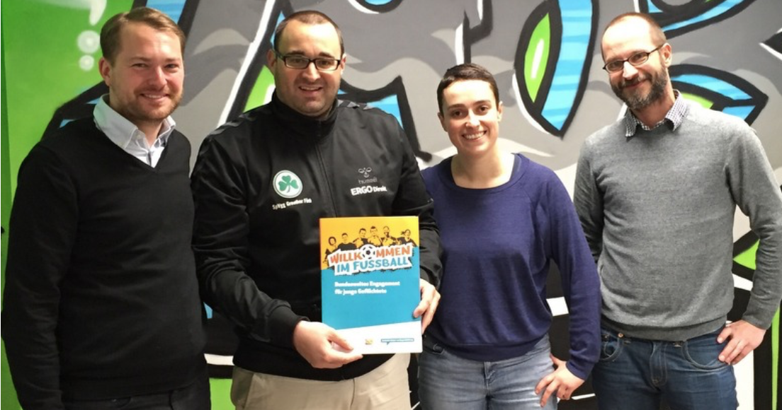 Vier Männer stehen lächelnd vor einer Wand mit Graffiti-Motiven. In der Mitte hält einer von ihnen ein Buch mit dem Titel "Willkommen im Fußball - Grundschule". Das Buch wird von der DFL Stiftung gefördert.