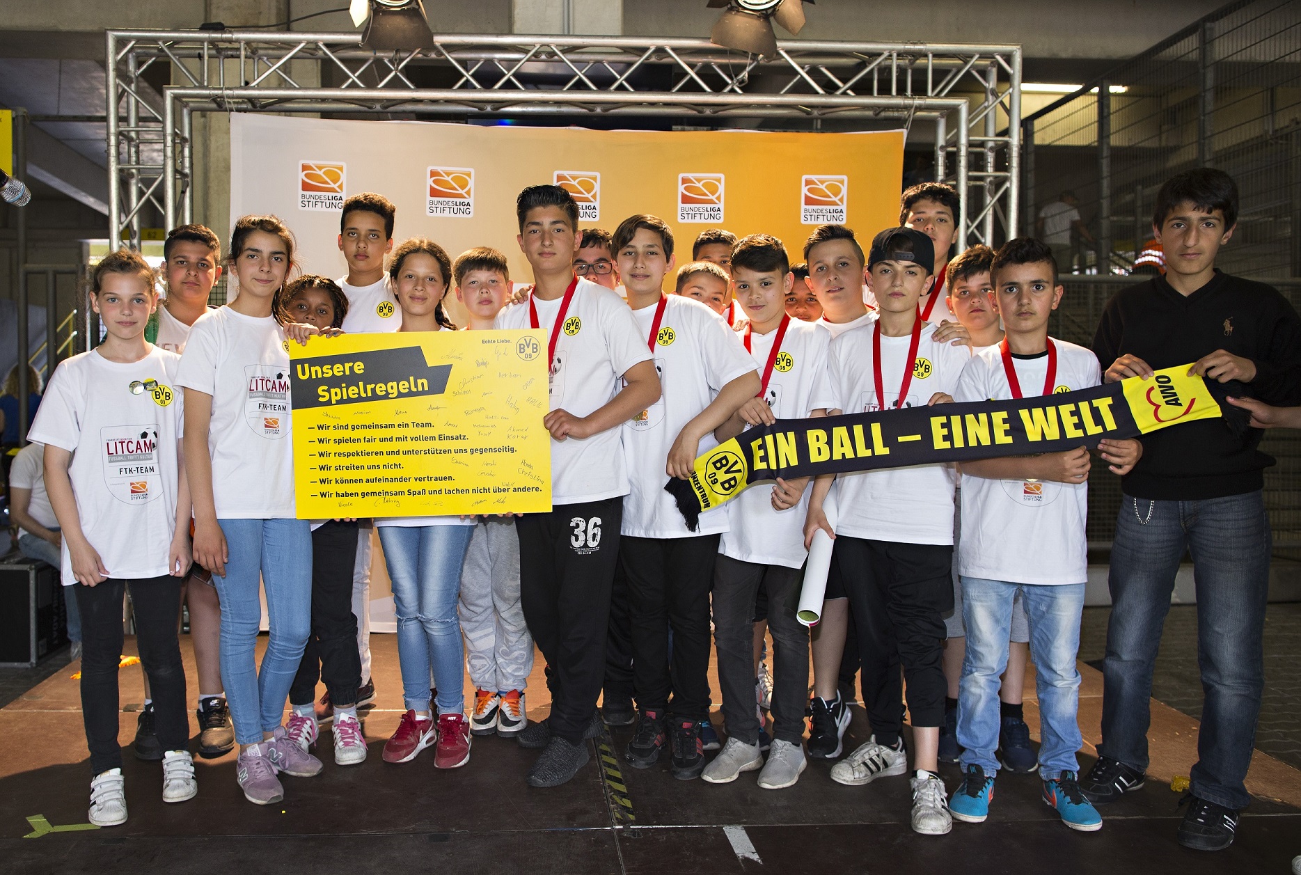 Eine Gruppe junger Fußballspieler steht lächelnd in einer Reihe vor einer Werbeplane mit dem Aufdruck "Ein Ball - eine Welt!". Im Hintergrund ist das Stadion zu erkennen.