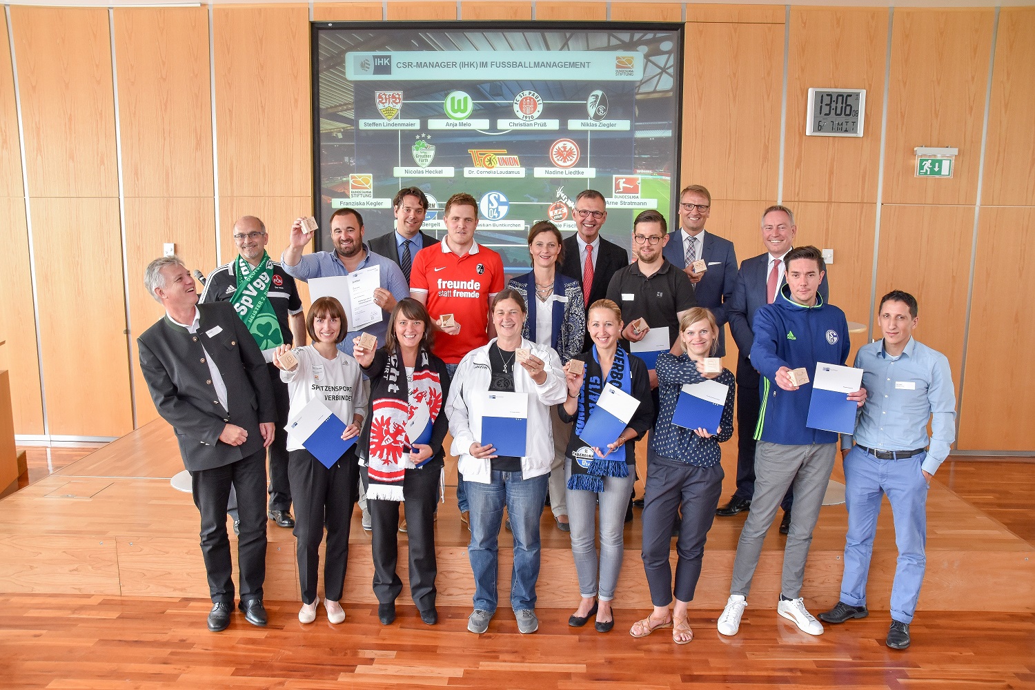 Eine Gruppe von rund 20 Personen steht in einem hellen Raum vor einer Wand mit Logos verschiedener Fußballvereine. Die Teilnehmer eines CSR-Managers-Programms im Fußball posieren für ein Gruppenfoto. Einige tragen Fußballschals und T-Shirts ihrer Vereine.