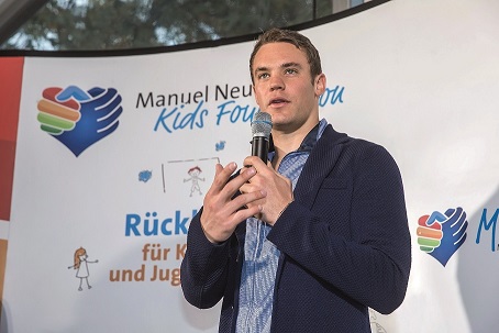 Manuel Neuer hält ein Mikrofon in der Hand und spricht auf einer Veranstaltung der DFL Stiftung. Im Hintergrund ist ein Banner mit dem Logo der Manuel Neuer Kids Foundation und dem Schriftzug "Rückhalt für Kinder und Jugend" zu sehen.