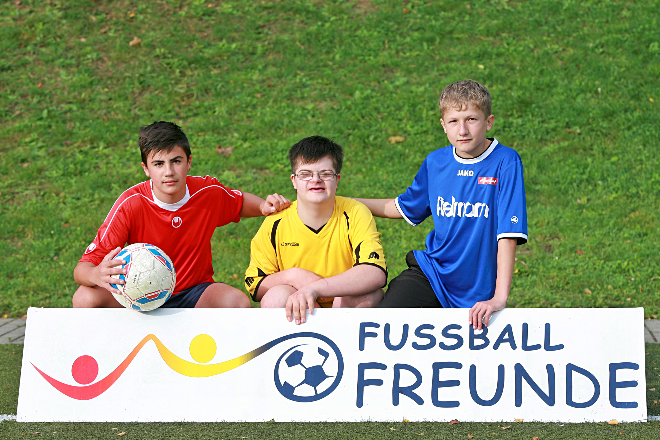 Drei Jugendliche, zwei mit und einer mit Down-Syndrom, sitzen nebeneinander auf einer Bank und lächeln in die Kamera. Sie tragen Trikots verschiedener Farben und halten einen Fußball. Im Hintergrund ein Banner mit der Aufschrift "Fußballfreunde".