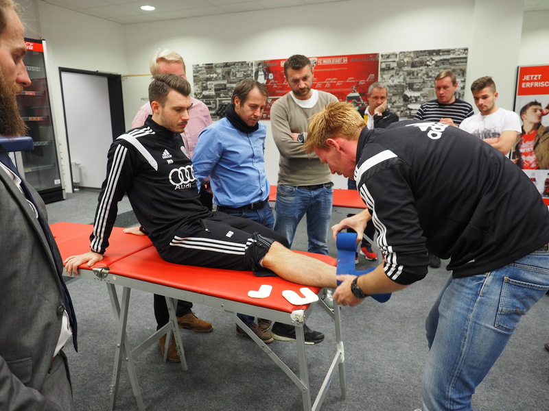 Fußballspieler liegt auf einer roten Bank, während ein Physiotherapeut sein Bein untersucht. Im Hintergrund stehen weitere Personen, vermutlich Teilnehmer einer Schulung.