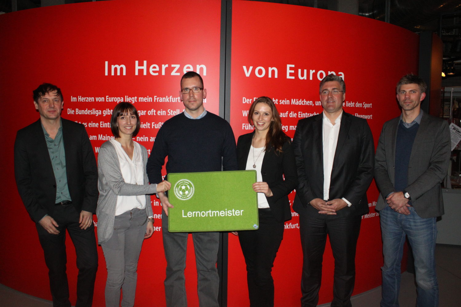 Eine Gruppe von sechs Männern und Frauen steht lächelnd vor einem Banner mit der Aufschrift "Im Herzen von Europa liegt mein Frankfurt". In den Händen halten sie eine grüne Tafel mit dem Logo einer Fußballschule und dem Schriftzug "Lerntmeister". Im Hintergrund sind weitere Banner mit Fußballbezogenen Texten zu sehen.