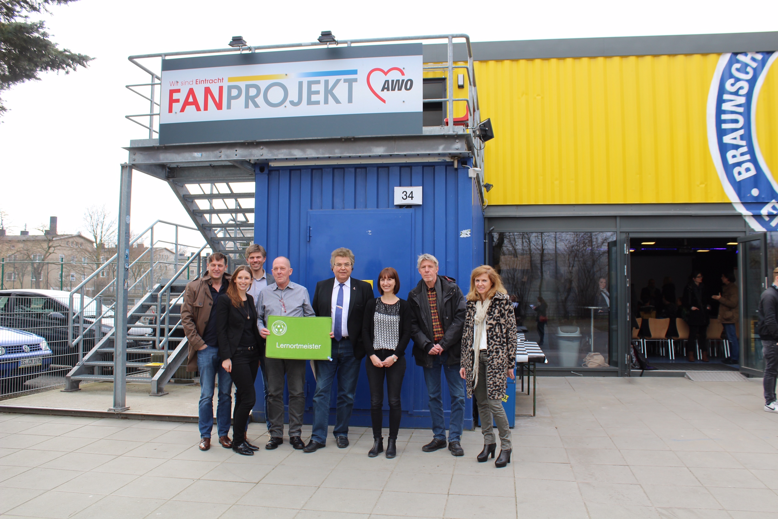 Eine Gruppe von Menschen steht vor einem blauen Container, der mit "FANPROJEKT" und einem roten Herzlogo beschriftet ist. Im Hintergrund ist ein gelber Bau mit der Aufschrift "Braunschweig" zu sehen.