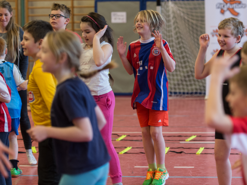 Eine Gruppe von Kindern und Jugendlichen nimmt an einer Bewegungseinheit in einer Sporthalle teil. Sie strecken und heben die Arme, während sie sich fröhlich bewegen. Im Hintergrund ist ein Fußballtor zu sehen.