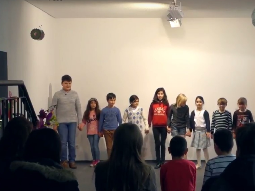 Eine Gruppe von Kindern und Jugendlichen steht auf einer Bühne und hält sich an den Händen. Im Hintergrund sind dunkelhaarige Zuschauer im Publikum zu erkennen.