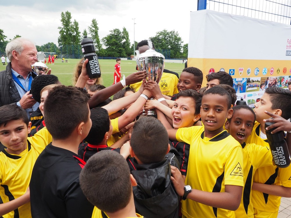 Jubelnde Kinder in gelben Trikots umarmen sich und heben einen Pokal in die Höhe. Im Hintergrund sind weitere Personen und ein Fußballfeld erkennbar.