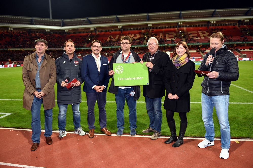 Eine Gruppe von sieben Personen steht auf der Laufbahn eines Fußballstadions. Sie halten eine grüne Tafel mit dem Aufdruck "Lernort Stadion" in der Hand. Im Hintergrund sind die Zuschauertribünen zu sehen.