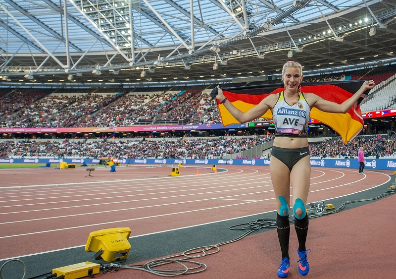Eine Athletin mit der deutschen Flagge über der Schulter steht auf einer roten Laufbahn in einem Stadion. Im Hintergrund ist ein volles Publikum zu sehen. Sie trägt Sportkleidung und hat einen jubelnden Gesichtsausdruck.