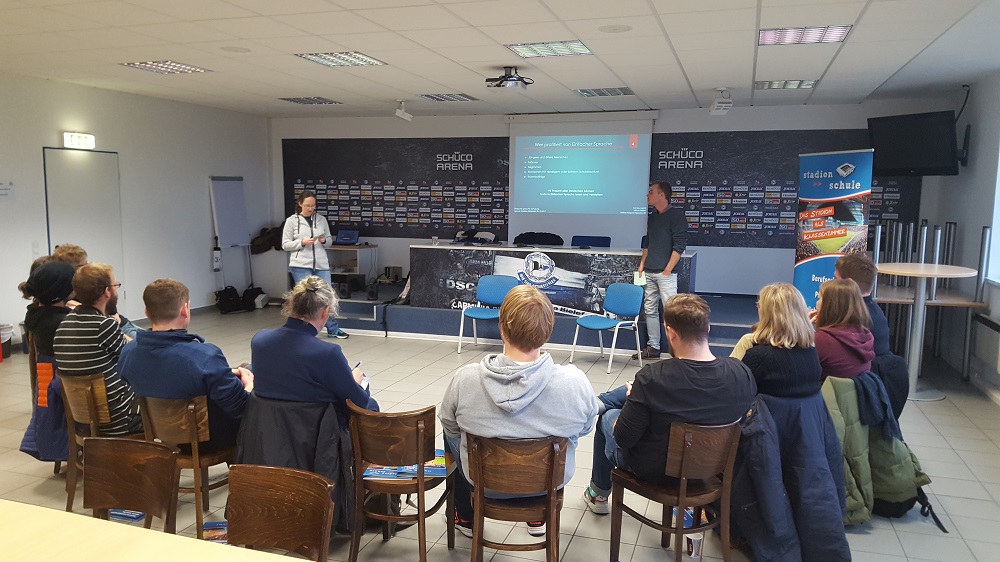 Eine Gruppe von Menschen sitzt in einem hellen Raum zusammen und nimmt an einer Präsentation teil. Vorne steht eine Person und spricht, während im Hintergrund eine Leinwand mit dem Schriftzug "SCHÜCO ARENA" und Informationen zu einem Lernort Stadion zu sehen ist.