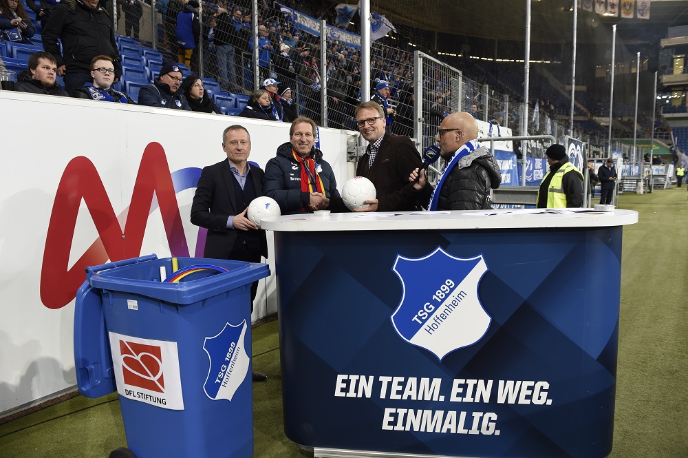 DFL Stiftung Logo auf einem Müllcontainer vor dem Stadion der TSG 1899 Hoffenheim bei einer Weihnachtsaktion. Im Hintergrund sind Personen und Sitzreihen erkennbar.