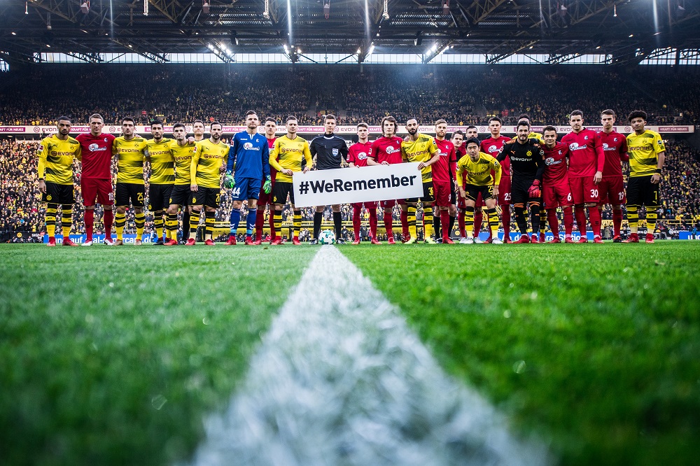 Fußballspieler von Borussia Dortmund und dem SC Freiburg stehen vor einem vollen Stadion und halten ein Banner mit dem Hashtag #WeRemember hoch. Im Hintergrund sind die Zuschauerreihen und die Flutlichtmasten erkennbar.