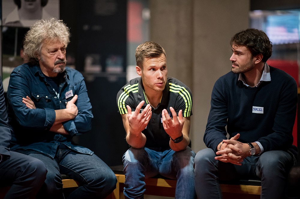 Drei Männer sitzen nebeneinander auf einer Bank in einer Stadion-Umgebung und diskutieren. Der Mann in der Mitte gestikuliert mit den Händen, während die beiden anderen ihm aufmerksam zuhören.