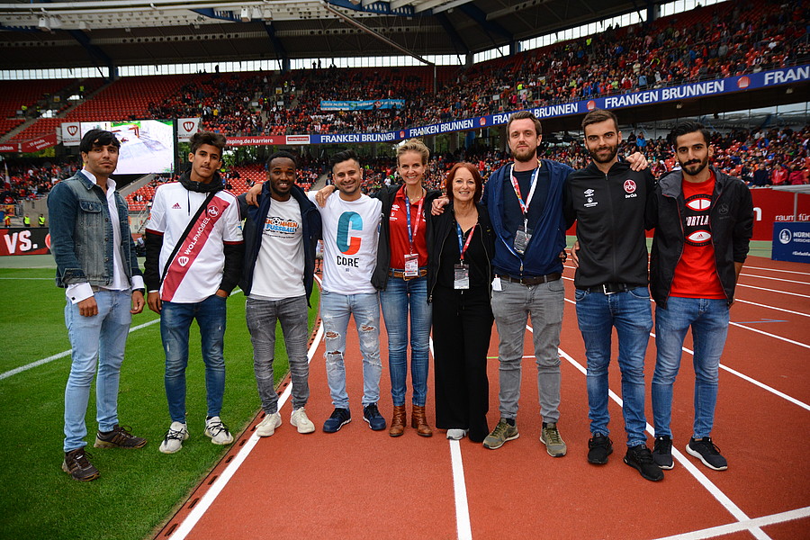 Eine Gruppe von jungen Männern und Frauen steht auf der roten Tartanbahn eines Fußballstadions. Die meisten tragen lässige Kleidung, einige haben rot-schwarze Schals oder T-Shirts des 1. FC Nürnberg um den Hals oder tragen sie. Im Hintergrund sind die leeren Zuschauerreihen des Stadions zu sehen.