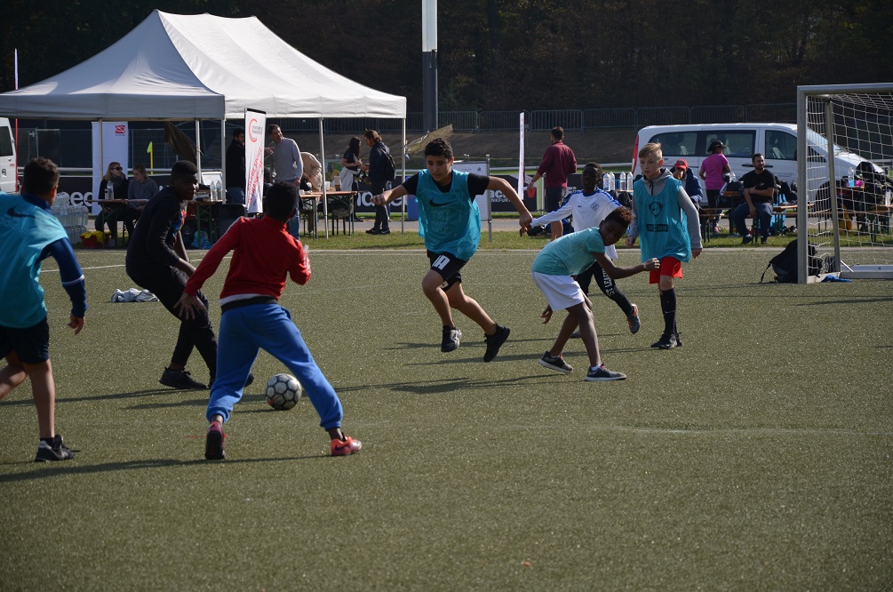 Eine Gruppe Jugendlicher spielt Fußball auf einem grünen Kunstrasenplatz. Einige Spieler sind in hellblauen Trikots, andere in Rot und Blau gekleidet. Im Hintergrund sind weitere Personen und Zelte zu sehen.