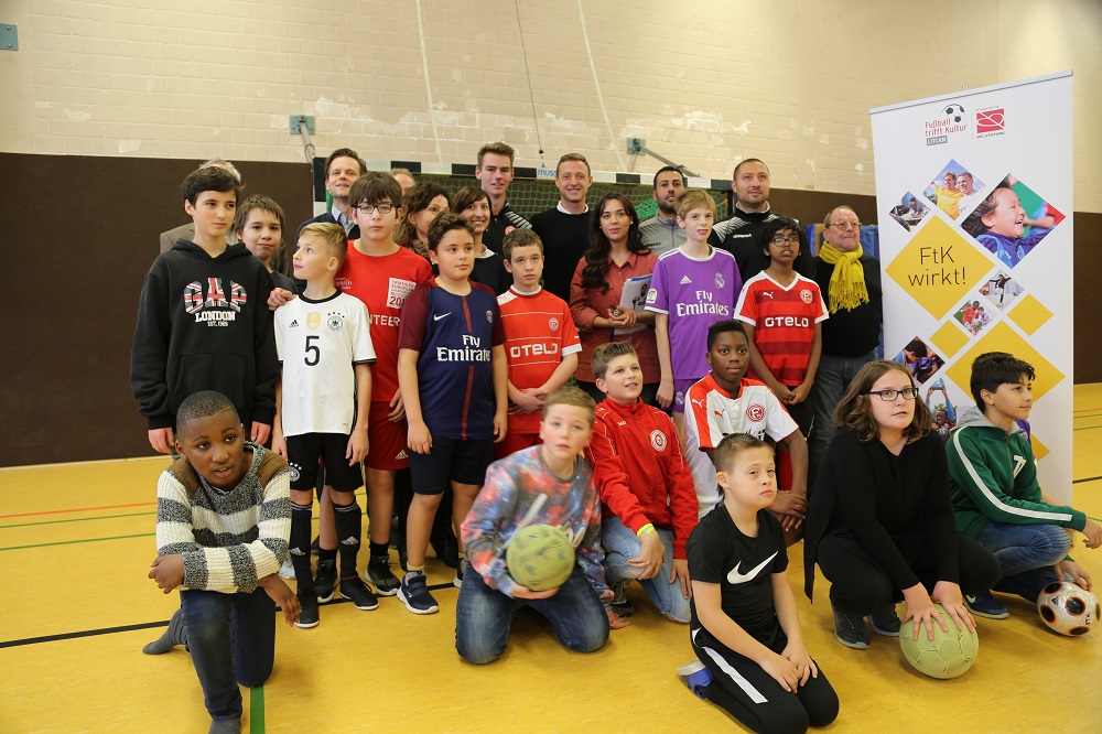 Eine Gruppe von Kindern und Jugendlichen steht in einer Turnhalle zusammen und lächelt in die Kamera. Viele tragen Fußballtrikots verschiedener Vereine. Im Hintergrund ein Banner mit der Aufschrift "FTK wirkt!" und dem Logo der DFL Stiftung.