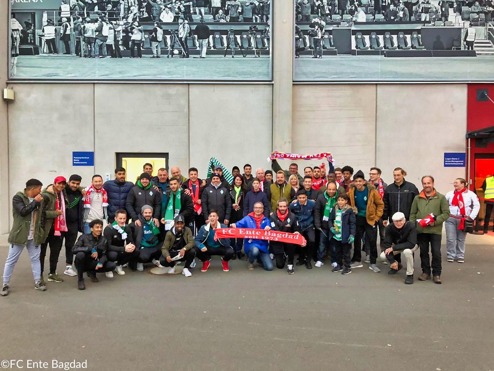 Eine große Gruppe von Menschen steht und kniet vor einer Stadionkulisse zusammen. Viele tragen Fanartikel des 1. FSV Mainz 05 und von Werder Bremen, was auf eine Fankultur-Veranstaltung oder ein Treffen von Fangruppen hindeutet. Im Vordergrund hält jemand eine Fahne mit der Aufschrift "FC Ente Bagdad".
