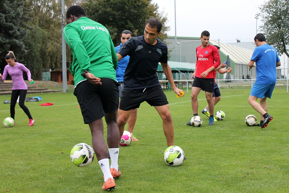 Fußballtraining mit Jugendlichen auf einem grünen Rasenplatz. Zwei dunkelhäutige Jugendliche üben Dribbling mit Fußbällen im Vordergrund, während im Hintergrund weitere Jugendliche in Trainingsanzügen ebenfalls mit Fußbällen agieren.