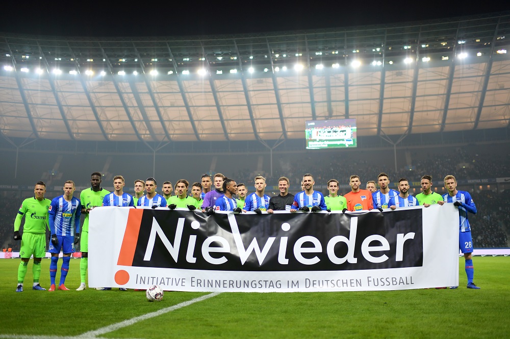 Mannschaften von Hertha BSC und Schalke 04 stehen mit einem Banner mit der Aufschrift "Niewieder" im Stadion. Das Banner ist Teil der Initiative zum Erinnerungstag im deutschen Fußball.
