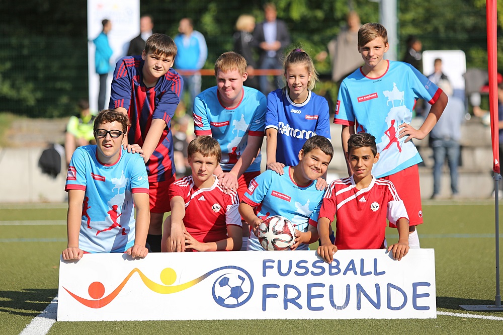 Eine Gruppe von Kindern und Jugendlichen in blauen und roten Fußballtrikots steht auf einem grünen Fußballplatz vor einem weißen Banner mit der Aufschrift "FußballFREUNDE" und dem Logo der Initiative.