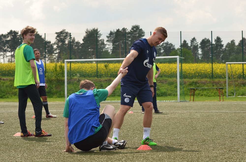 Ein Fußballspieler im blauen Trainingsanzug hilft einem anderen Spieler, der auf dem Boden sitzt, auf die Beine. Im Hintergrund stehen weitere Spieler in Trainingskleidung auf einem Fußballfeld.