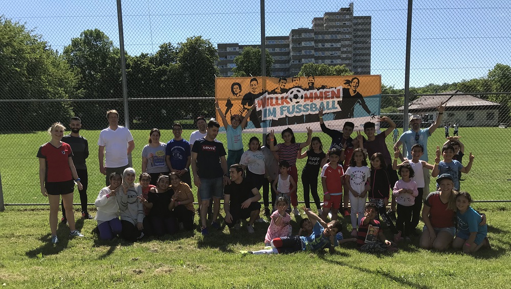 Eine große Gruppe von Menschen, darunter viele Kinder, steht auf einem Fußballfeld und lächelt in die Kamera. Im Hintergrund ist ein Banner mit der Aufschrift "Willkommen im Fußball" zu sehen. Die Szene zeigt ein Ballsportturnier für Geflüchtete in Stuttgart, das von der DFL Stiftung unterstützt wird.