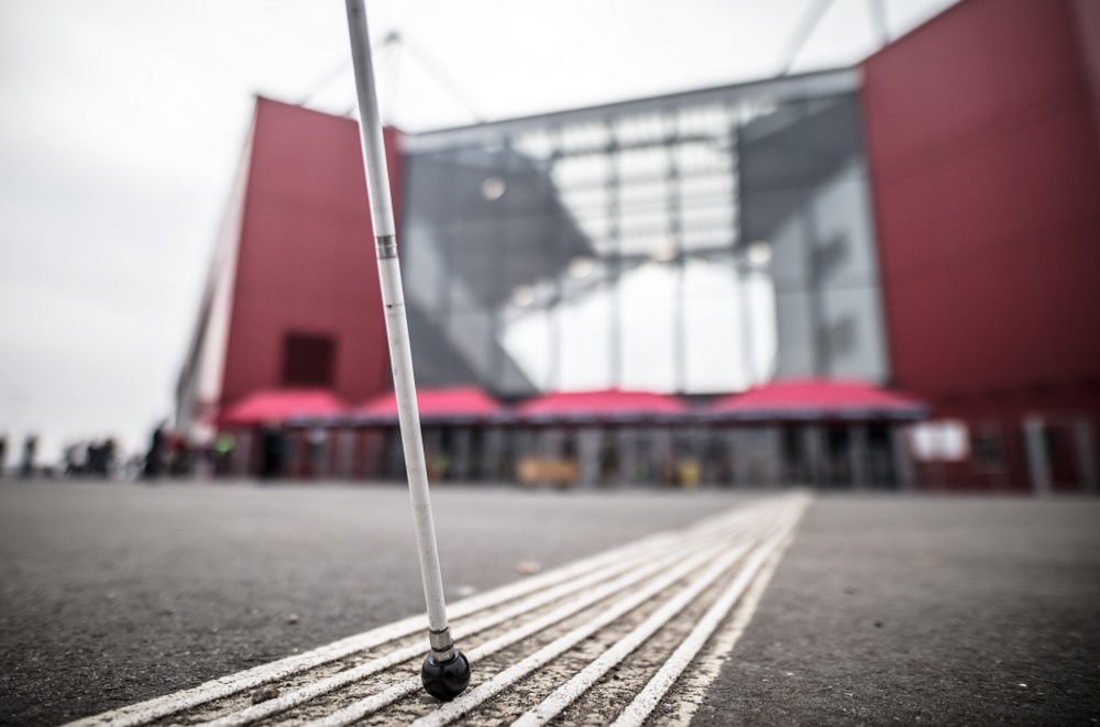 Ein blinder Gehstock berührt eine tactile Leitspur vor dem Stadion, um blinden und sehbehinderten Fans den Weg zu weisen.