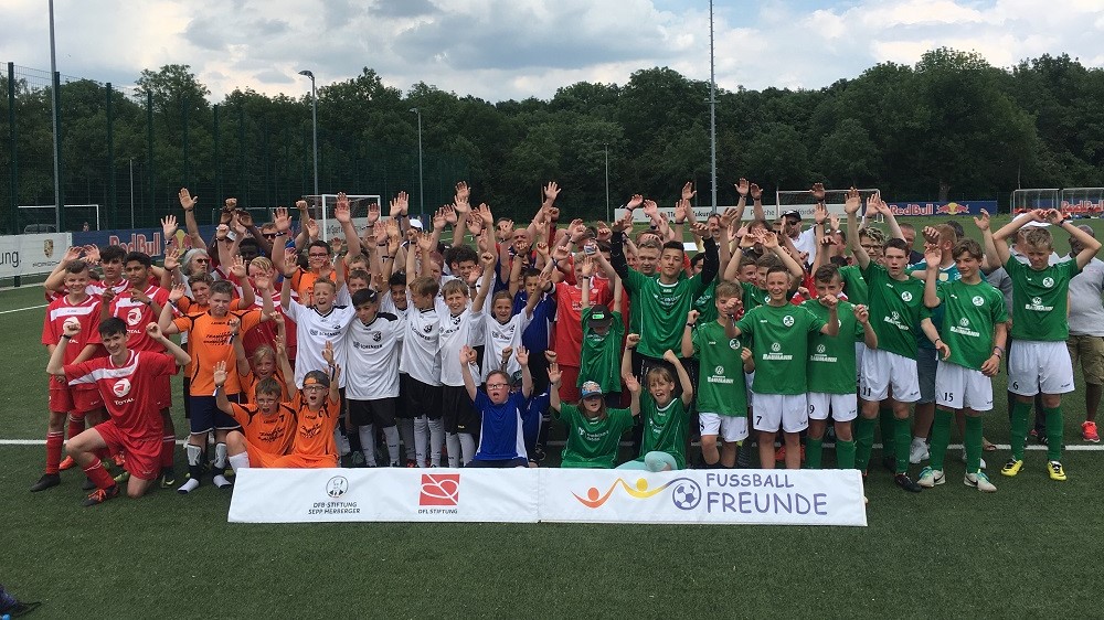 Eine Gruppe von Kindern und Jugendlichen in Fußballtrikots steht auf einem grünen Fußballfeld und lächelt in die Kamera. Links tragen einige rote Trikots, während die Mehrheit grüne Trikots mit dem Schriftzug "DFL" trägt. Im Hintergrund sind Bandenwerbung und ein Fußballtor zu sehen. Ein Banner mit dem Logo "Fußballfreunde" liegt vor der Gruppe.