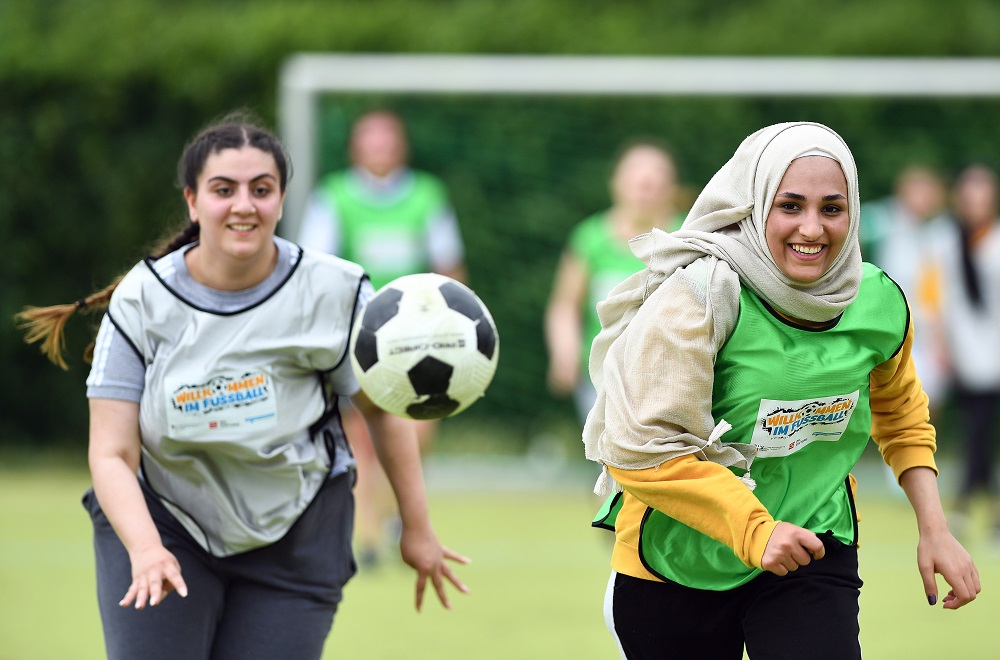 Zwei junge Frauen mit Kopftuch spielen auf einer grünen Wiese Fußball. Die Frau im Vordergrund in grünem T-Shirt und schwarzer Hose lächelt und scheint gerade einen Pass zu spielen. Die zweite Frau im roten T-Shirt versucht den Ball mit dem Kopf zu kontrollieren. Im Hintergrund sind weitere Personen erkennbar.