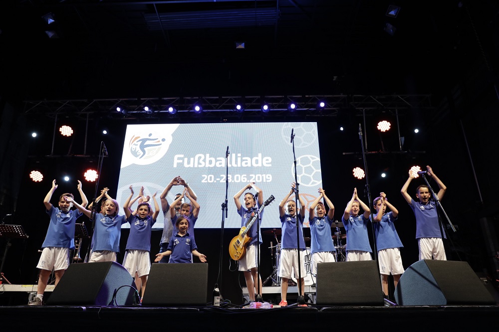 Gruppe von Kindern und Jugendlichen mit blauen T-Shirts und weißen Shorts auf einer Bühne, die mit erhobenen Händen jubeln. Im Hintergrund eine Leinwand mit der Aufschrift "Fußballiade 20. - 23.06.2019".