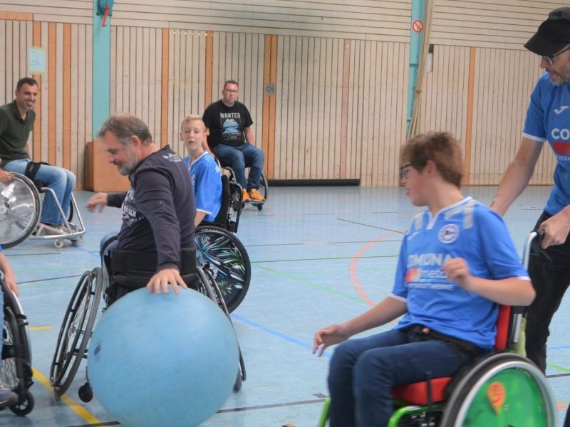 Rollstuhl-Fußballturnier der RBG Dortmund 51: Mehrere Spieler im Rollstuhl auf dem Spielfeld, einige konzentrieren sich auf den blauen Ball, während andere pausieren oder sich austauschen. Im Hintergrund sind weitere Personen und die Hallenwand zu sehen.