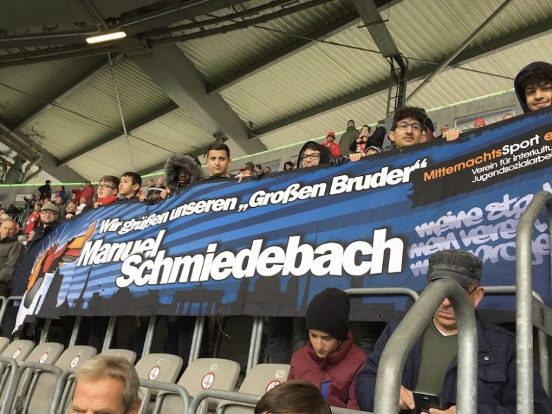 Banner mit der Aufschrift "Wir grüßen unseren ‚Großen Bruder‘ Manuel Schmiedebach" im Stadion während eines Fußballspiels. Im Hintergrund sind Zuschauerreihen zu sehen.