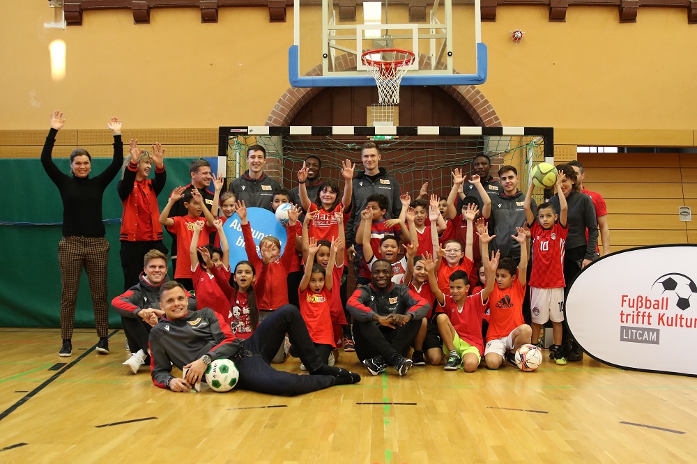 Gruppe von Kindern und Jugendlichen in roten Trikots, umgeben von Betreuern und DFL Stiftung Mitarbeitern, jubelt in einer Sporthalle. Im Hintergrund ein Basketballkorb und ein Banner mit dem Aufdruck "Fußball trifft Kultur LITCAM".