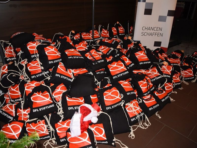 Stapelweise rote Schals mit dem Aufdruck "DFL STIFTUNG" liegen auf einem Tisch, vermutlich als Gastgeschenke bei einem Weihnachtsessen. Im Hintergrund ist ein Schild mit der Aufschrift "CHANCEN SCHAFFEN" zu sehen.