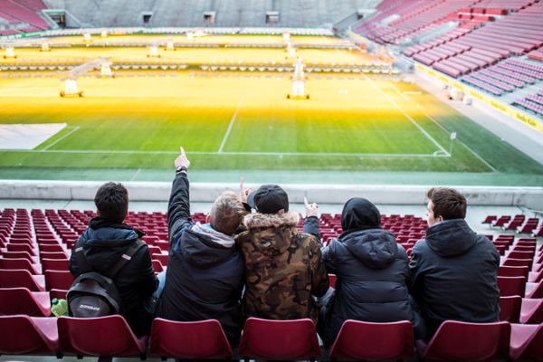 A small group of students sits together in an empty soccer stadium and looks out onto the field from the stands. The group is taking part in the DFL Foundation's