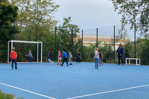 Mehrere Kinder spielen auf einem Bolzplatz mit einem blauen Bodenbelag gemeinsam Fußball.