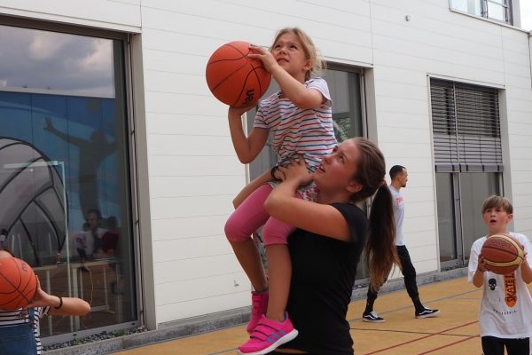 Ein Mädchen sitzt auf der Schulter einer jungen Frau und hält einen Basketball in der Hand. Die junge Frau hilft ihr dabei, den Ball in den Korb zu werfen.