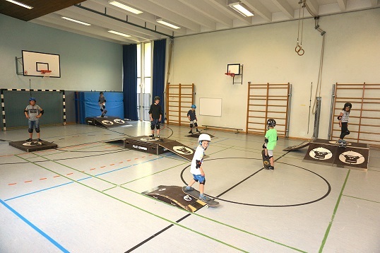 In einer Turnhalle sind verschiedene Rampen aufgebaut, die von Kindern befahren werden, die Helme und Schoner tragen.